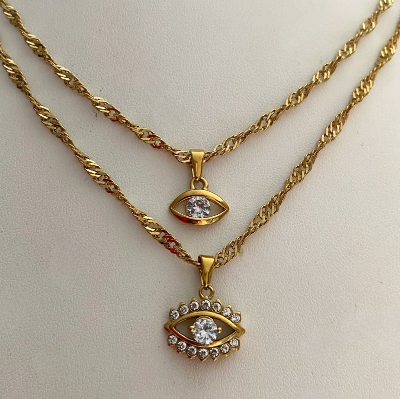 GRISÉ NYC layered gold evil eye pendant necklaces featuring twisted chains, sparkling cubic zirconia accents, and 18k gold-plated stainless steel construction.