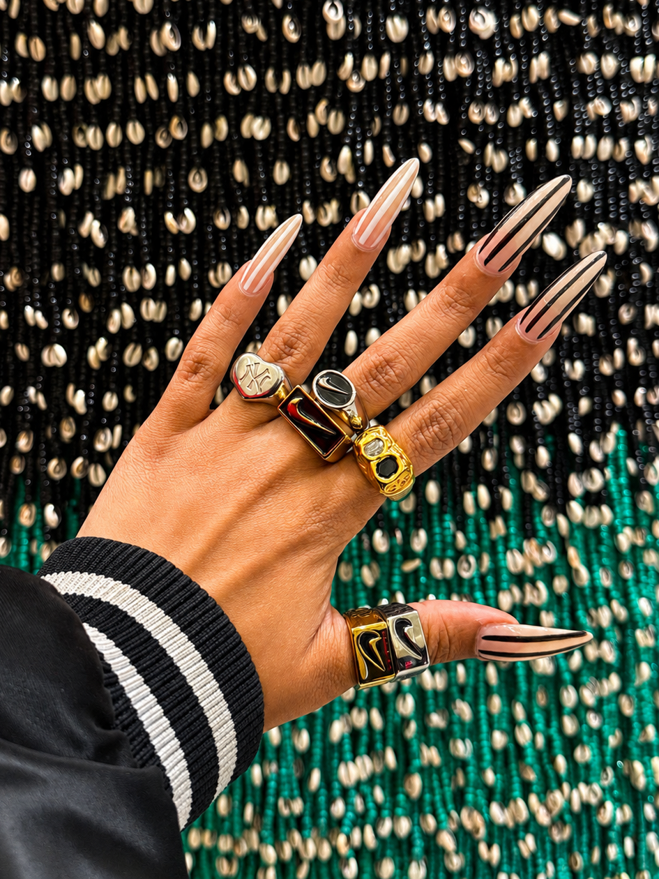 Close-up of stacked Just Do It stainless steel rings worn with statement jewelry against a textured backdrop.