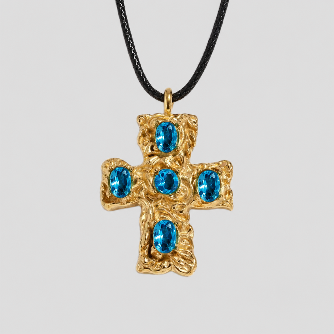 GRISÉ NYC Blue gemstone cross pendant necklace in gold with five oval blue stones on a black cord necklace against a light gray background.