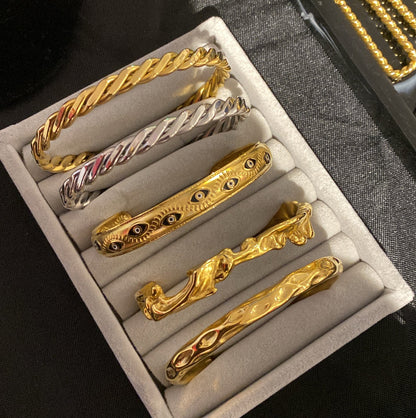 GRISÉ NYC 18k gold-plated Evil Eye Enamel Cuff Bracelet displayed alongside a collection of gold bracelets in a grey jewelry organizer.