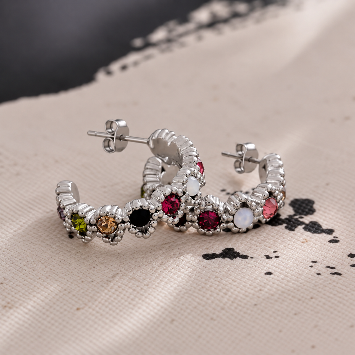 GRISÉ NYC Gemma Multicolor Silver Hoop Earrings featuring colorful cubic zirconia stones and butterfly backs, styled on textured beige fabric.