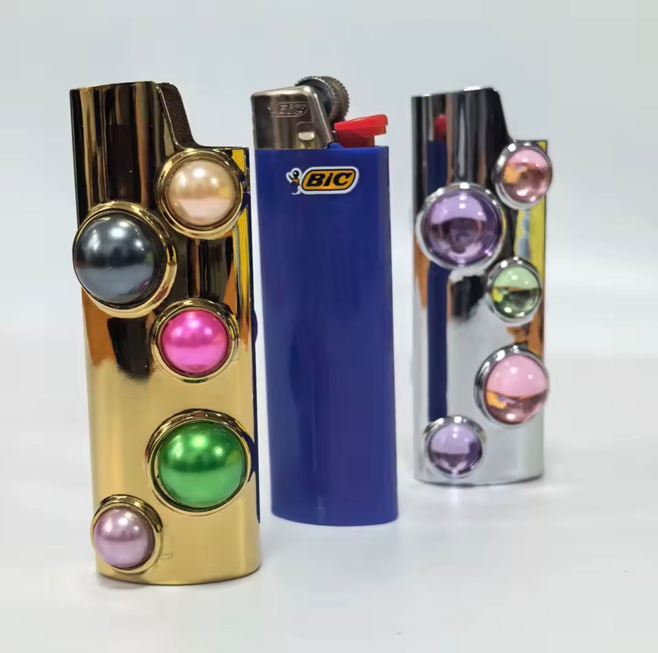Luxury gold and silver gemstone lighter case collection by GRISÉ NYC with colorful cabochon stones, shown alongside a blue BIC lighter for size reference.
