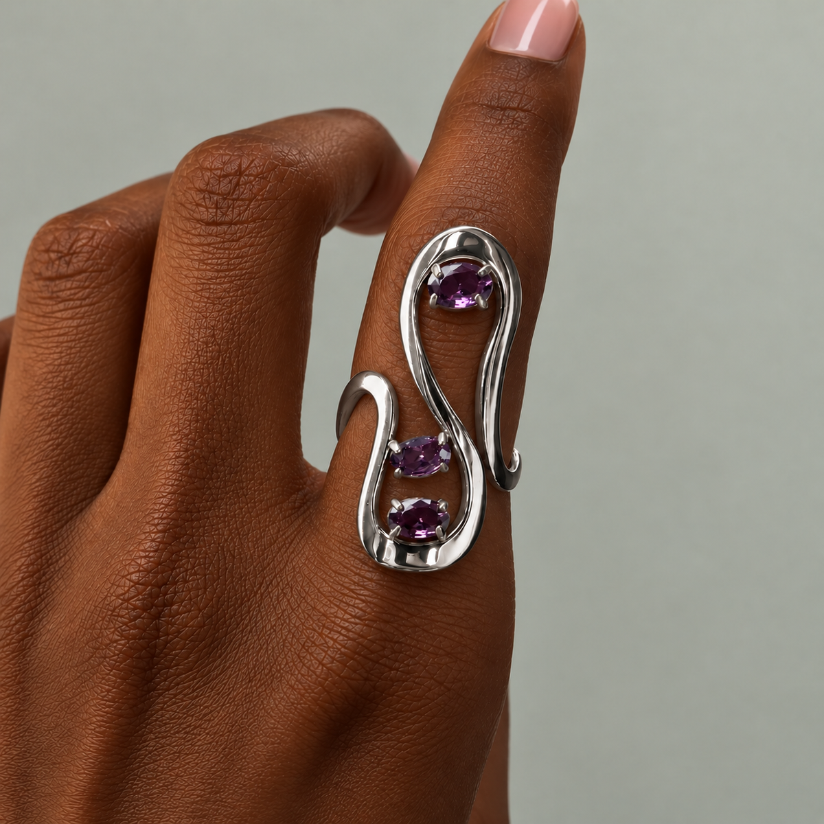 Close-up of a sculptural silver statement ring featuring three oval purple gemstones in a flowing, open-wrap design, styled on hand with a nude manicure.