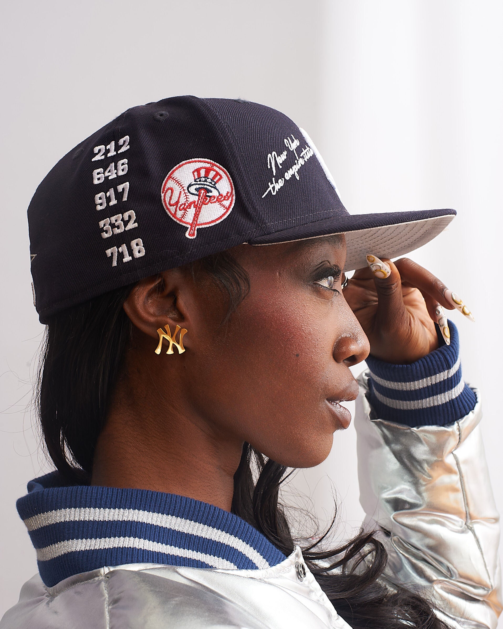 Side profile of female model wearing GRISÉ NYC gold New York NY logo stud earrings styled with black cap modern unisex streetwear look.