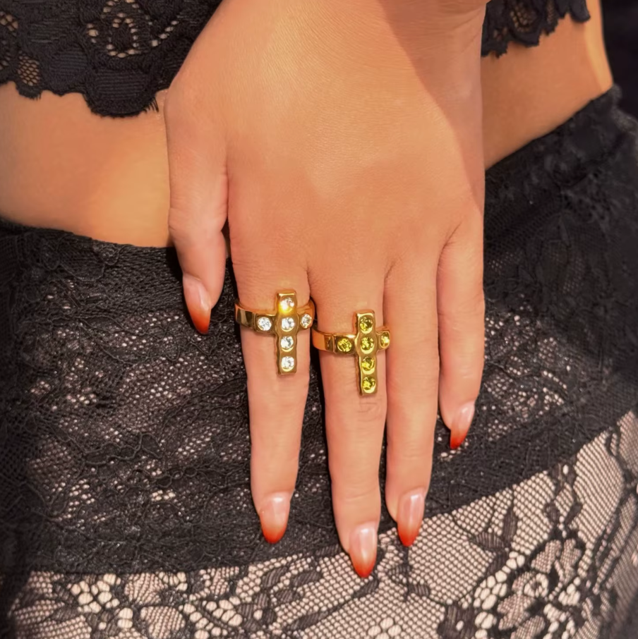 Two Saint Ice Cross 18k Gold Rings by GRISÉ NYC with clear and green crystal accents, stacked and styled on hand in natural sunlight.